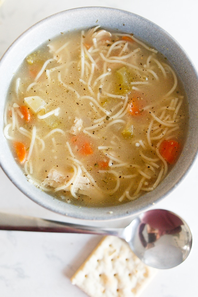 A grey bowl full of homemade chicken noodle soup. You can see carrots and noodles in the broth. A spoon with a cracker is below the bowl.